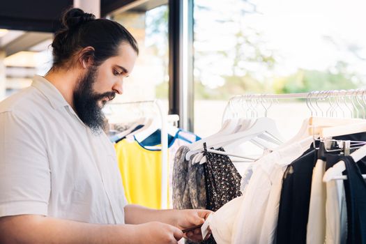 young man with beard, looking and buying clothes, in fashion shop. shopping concept. leisure concept. Natural light, sunbeams, clothes rack with coloured clothes, horizontal view, copy space. dress with white shirt.
