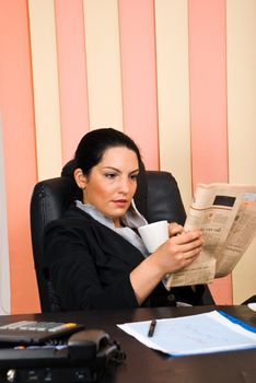 Young business woman sitting comfortable in her chair at office,drinking a cup of coffee and reading a newspaper