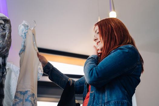 Young woman with red hair, looking at a T-shirt to buy, in a fashion shop. concept of shopping. concept of leisure. Natural light, sun rays, display with clothes, clothes rack, customers in background walking, clothes, vertical view. customer wearing blue denim jacket, red t-shirt and jeans.