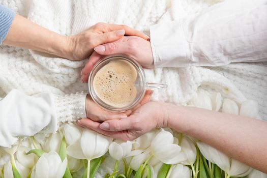 four hands hold one cup of heart-shaped cappuccino, floral background with white tulips, drink coffee in the company, female friendship, lgbt. High quality photo