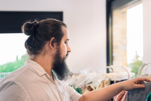 young man with beard, in fashion shop, buying a t-shirt as a present for his girlfriend. shopping concept. romantic concept. Natural light, sunbeams, clothes rack with coloured clothes, horizontal view, copy space. dress with white shirt.