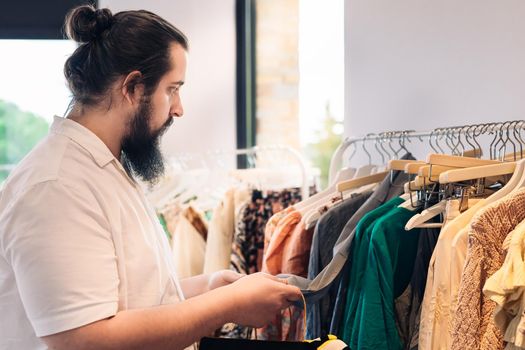 young man with beard, in fashion shop, buying a t-shirt as a present for his girlfriend. shopping concept. leisure concept. Natural light, sunbeams, clothes rack with coloured clothes, horizontal view, copy space. dress with white shirt.
