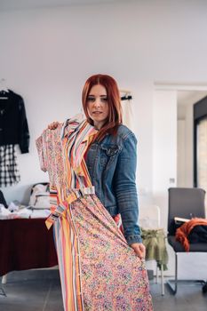 young woman trying on a dress in front of a mirror. red-haired girl shopping in a fashion shop. shopping concept. leisure concept. Natural light, sunbeams, display, clothes rack, clothes, vertical view, space to copy. customer in blue denim jacket, red t-shirt.