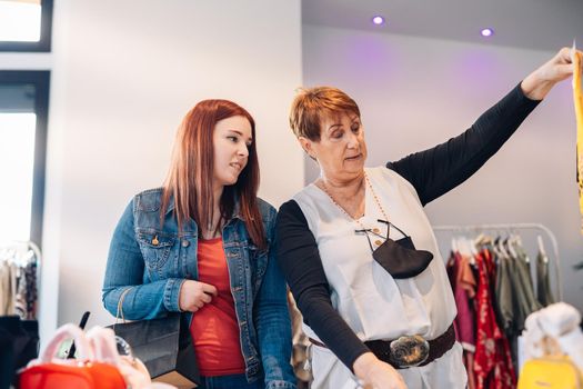 old woman and young woman buying clothes in a fashion shop. grandmother and granddaughter enjoying a shopping day. shopping concept. leisure concept. two smiling and happy people. Natural light, sunshine, clothes rack with colourful clothes, horizontal view, space for copying. casually dressed.