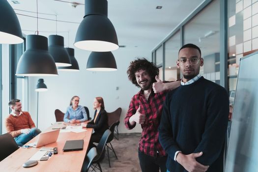 Portrait of two happy millennial male business owners in modern office. Two businessmen smiling and looking at the camera. Busy diverse team working in the background. Leadership concept. Head shot. High-quality photo