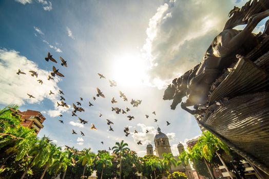 Horizontal general view of Bolivar park located in Pereira Risaralda, with many birds flying next to the monument the naked bolivar and sun in the clear sky