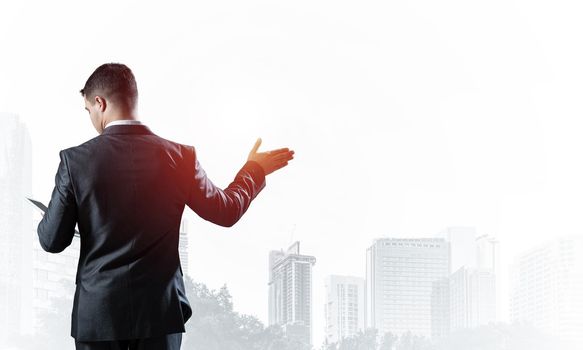 Handsome businessman standing and looking into documents. Back view of man in business suit and tie on background of foggy cityscape. Successful business person with raised hand and open palm.