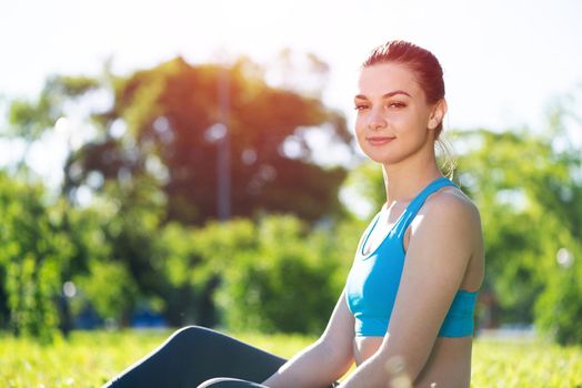 Beautiful smiling girl in sportswear relax in park. Young woman with sitting on green grass after training. Meditation outdoor at sunny summer day. Morning exercises and healthy lifestyle.