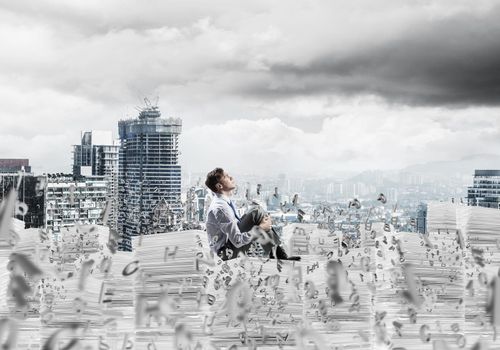 Young businessman looking away while sitting on pile of documents among flying letters with cityscape on background. Mixed media.