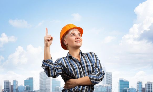 Beautiful female builder in hardhat with finger pointing upward. Portrait of smiling young woman in checkered blue shirt standing on background of cityscape. Industrial architecture and construction