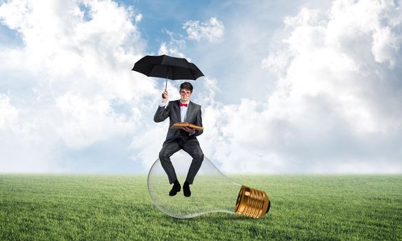 young businessman with an umbrella and a book, sitting on a light bulb. concept of finding new ideas in business