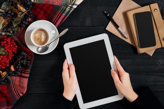 Women hands holding the tablet with black screen above the table with a cup of coffee and smart. Top view, flat lay. Copy space. Still life. Cafe.