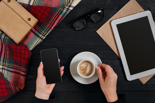 Women hands holding the phone with black screen above the table with a cup of coffee and tablet. Top view, flat lay. Copy space. Still life. Cafe.