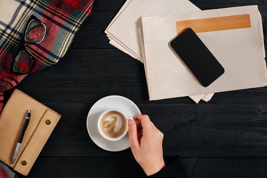 Newspaper and smartphone, reading glasses, notebook, hands holding coffee cup on a black wooden background. Stylish office desk. Autumn or Winter concept. Flat lay, top view. Still life. Copy space