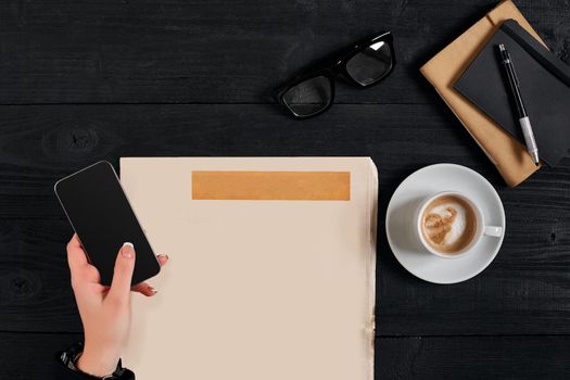 Newspaper and coffee cup, reading glasses, notebook, hands holding smartphone on a black wooden background. Stylish office desk. Autumn or Winter concept. Flat lay, top view. Still life. Copy space