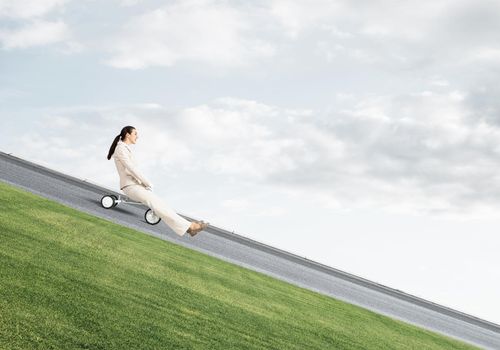 Beautiful happy woman riding downhill on road. Young employee in white business suit going down on small bike outdoor. Fast business startup. Beginner level business concept with bicyclist
