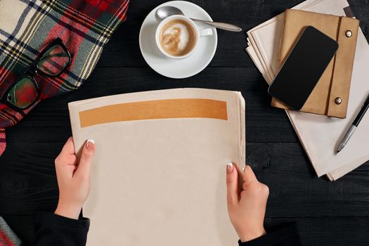 Smartphone and coffee cup, reading glasses, notebook, hands holding newspaper on a black wooden background. Stylish office desk. Autumn or Winter concept. Flat lay, top view. Still life. Copy space