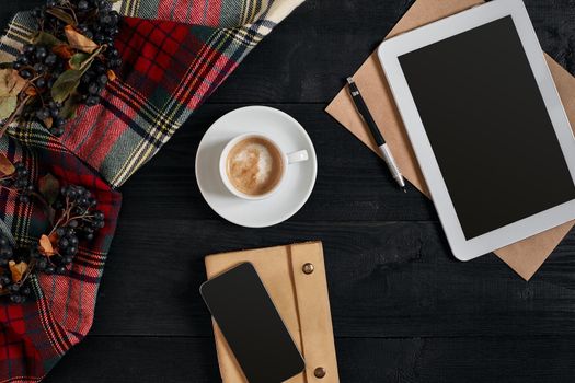 Above view of Smart phone, tablet, scarf in a cage, glasses with notebook and cup of latte coffee on black wooden background. Still life. Copy space. Flat lay. Top view