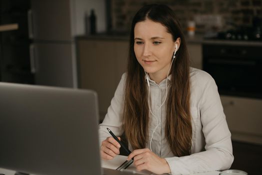 Remote work. A caucasian brunette woman with headphones working remotely online on her laptop. A girl in a white shirt doing a video call to her business partners at her home workplace.