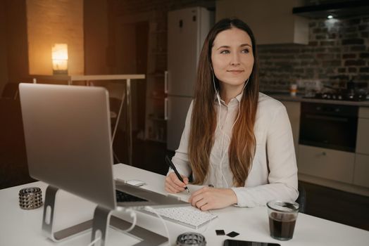 Remote work. A caucasian woman with headphones working remotely on her laptop. A happy brunette girl with a smile doing notes during an online business briefing at her cozy home workplace.