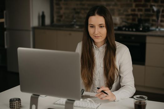 Remote work. A caucasian brunette woman with headphones working remotely on her laptop. A lady in a white shirt doing business at her home workplace.