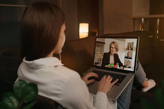 Back view of a woman lying at home on a sofa talking with her boss and other colleagues in a video call on a laptop. Businesswoman talks with coworkers on a webcam conference. A team having a meeting.
