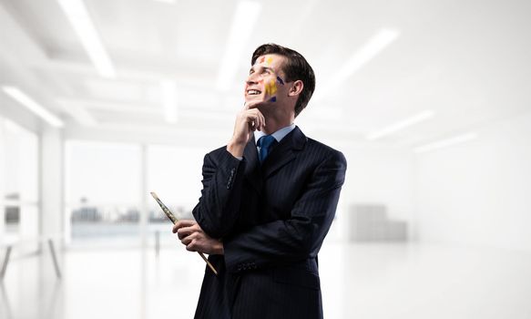 Portrait of young thoughtful man holding paintbrush in his hand and looking away while standing inside modern office view on background.