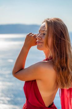 Smiling young woman in a red dress looks at the camera. A beautiful tanned girl enjoys her summer holidays at the sea. Portrait of a stylish carefree woman laughing at the ocean