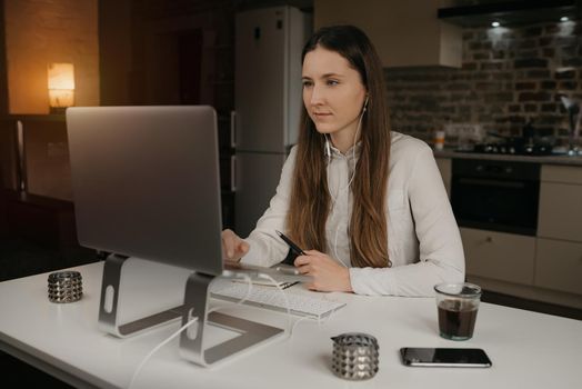 Remote work. A caucasian brunette woman with headphones working remotely online on her laptop. A girl in a white shirt doing a video call to her business partners at her cozy home workplace.