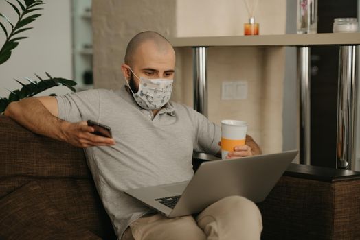 A man in a face mask working remotely on his laptop during the quarantine to avoid the spread coronavirus. A guy works from home holding a phone and a cup of tea during the pandemic of COVID-19.