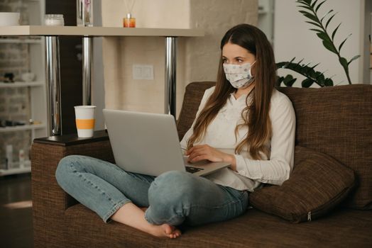 Remote work. Woman in a medical face mask studying remotely on her laptop during the quarantine to avoid the spread coronavirus. A girl working from home in a face mask during the pandemic of COVID-19