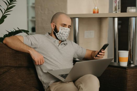 Remote work. Man in a medical face mask working remotely on his laptop during the quarantine to avoid the spread coronavirus. A guy works from home holding a smartphone during the pandemic of COVID-19