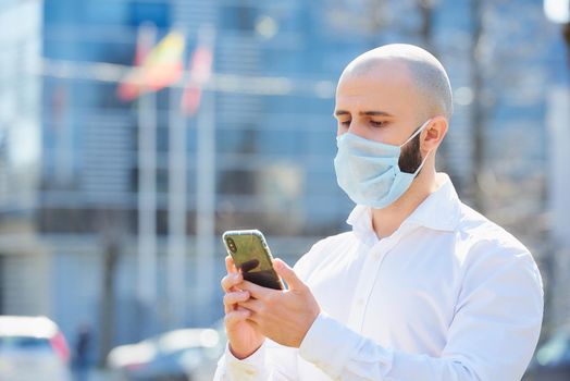 A bald man doing business with a smartphone wears a medical face mask to avoid the spread of coronavirus in the street. A close-up photo of a fellow with a surgical mask on the face against COVID-19.