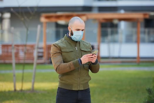 A bald man in a medical face mask to avoid the spread coronavirus holding his smartphone in the cozy street. A guy with a beard wears a face mask against COVID 19.