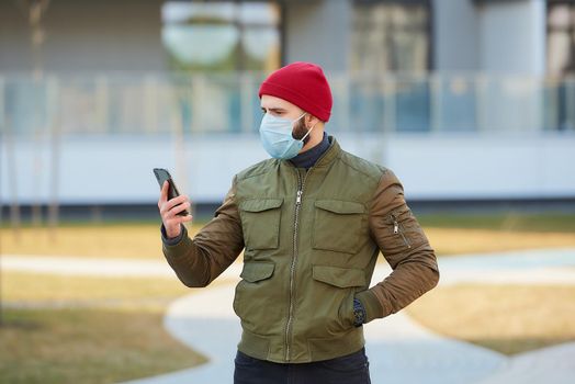 A man in a medical face mask to avoid the spread coronavirus checking his smartphone in the cozy street. A guy standing turned sideways wears a red cap and a face mask against COVID 19.