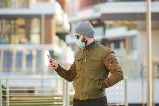 A man in a medical face mask to avoid the spread coronavirus checking his smartphone in the cozy street. A guy standing turned sideways wears a gray cap, sunglasses, and a face mask against COVID 19.