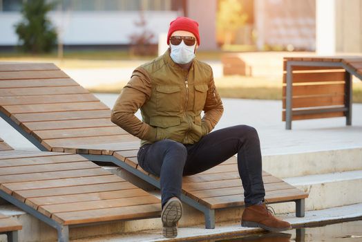 A man in a medical face mask to avoid the spread coronavirus in the cozy street. A guy sitting on a wooden deck chair dropping his hands in pockets wears sunglasses and a face mask against COVID 19.