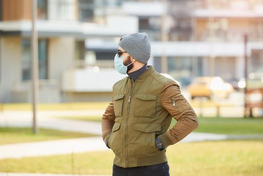 A man in a medical face mask to avoid the spread coronavirus waiting in the cozy street. A guy dropping his hands in his pockets wears a cap, sunglasses, and a face mask against COVID 19.