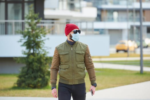 A man in a medical face mask to avoid the spread coronavirus holding his smartphone in a cozy street. A guy looking around wears a red cap, sunglasses, and a face mask against COVID 19.