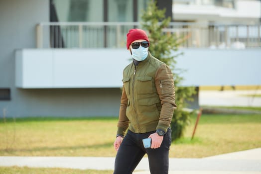 A man in a medical face mask to avoid the spread coronavirus holding his smartphone in a cozy street. A guy parading wears a red cap, sunglasses, and a face mask against COVID 19.