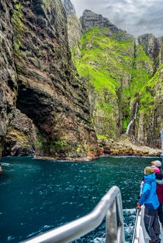 Blurred tourists observe the Vestmanna steep cliffs and natural stone bridge from the boat in Faroe Islands
