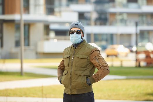 A man in a medical face mask to avoid the spread coronavirus waiting in the cozy street. A guy dropping his hands in his pockets wears a cap, sunglasses, and a face mask against COVID 19.