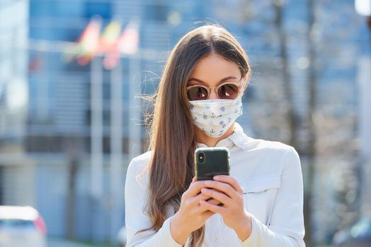 A woman doing business with a smartphone wears a medical face mask to avoid the spread of coronavirus in the street. A close-up photo of a lady with a surgical mask on the face against COVID-19..