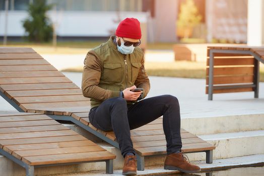 A man in a medical face mask to avoid the spread coronavirus using a smartphone in the cozy street. A guy sitting on a wooden deck chair wears sunglasses and a face mask against COVID 19.