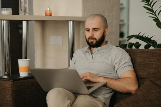 Remote work. A man working remotely on his laptop. A bald businessman with a beard works sitting on a sofa at home.