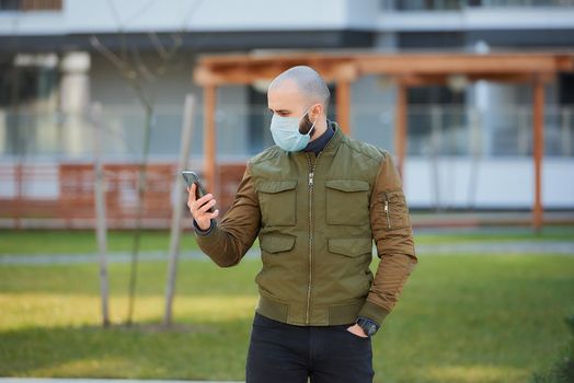 A bald man in a medical face mask to avoid the spread coronavirus checking his smartphone in the cozy street. A guy with a beard standing turned sideways wears a face mask against COVID 19.