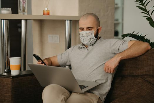 Remote work. A man in a medical face mask working remotely on his laptop during the quarantine to avoid the spread coronavirus. A guy works from home holding a phone during the pandemic of COVID-19.