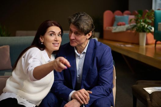 Handsome Caucasian man looking at his wife who is pointing on home goods in furniture shopping mall. Charming European woman with her husband looking for new sofa. Home improvement, interior design