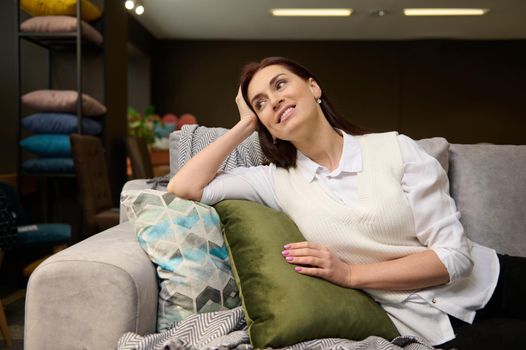 Charming European woman relaxing on a new comfortable sofa, testing it before buying it. Attractive woman buys a new stylish sofa in an upholstered furniture mall. Consumerism, interior design concept