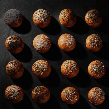 Assortment of fresh, delicious baked buns with black and white sesame and sunflower seeds against black background with copy space. Rural cuisine or bakery. Close-up, top view